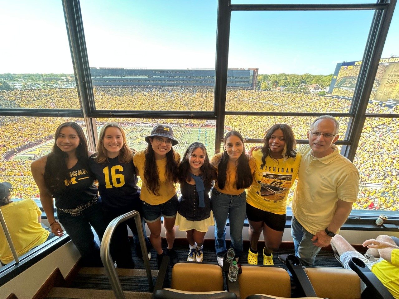 Group of 7 students and alum with their arms around each other  at a University of Michigan game with the crowd and field in the background 