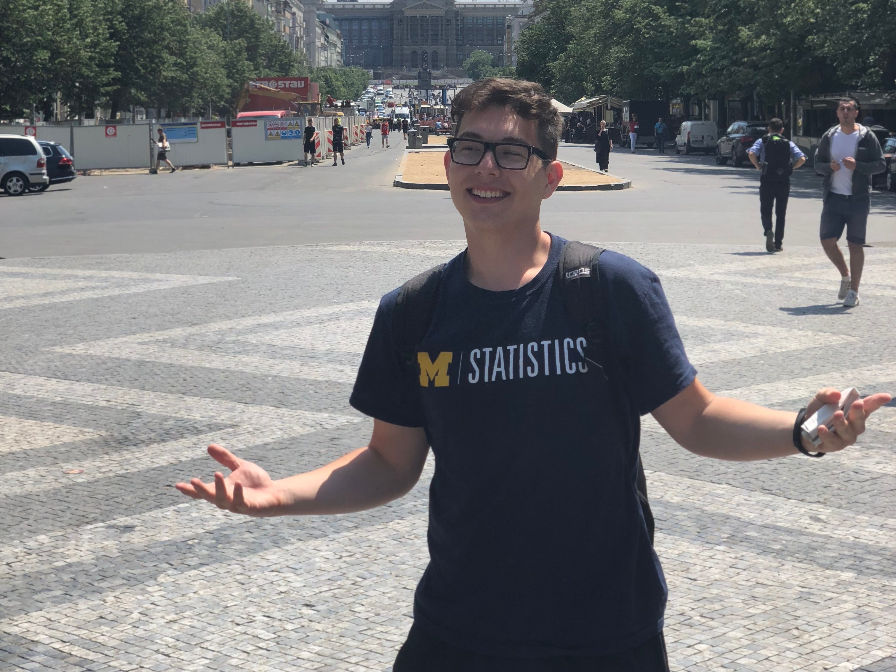 A student with his arms outstretched, grinning, stands in the center of a plaza wearing a University of Michigan Statistics shirt.