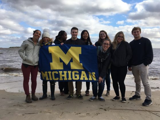 Students hold up a University of Michigan flag while standing on a beach with their backs to the water.