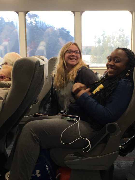In their seats on a train, two students smile for a photo. The student who is farthest from the camera sits up, while the other one reclines back.