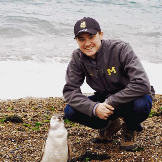 Alexander stands with his back to the sea, posing next to a bird. He's sporting a baseball cap and a grey zip-up shirt with an embroidered block M on his left chest.