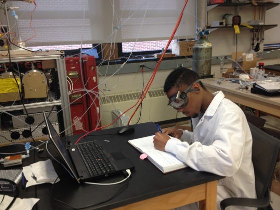 Black student in safety glasses and lab coat at work at a table in a lab