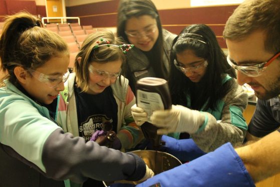 girl putting chocolate syrup in a bowl where liquid nitrogen is steaming off while a group of girls watches with graduate student