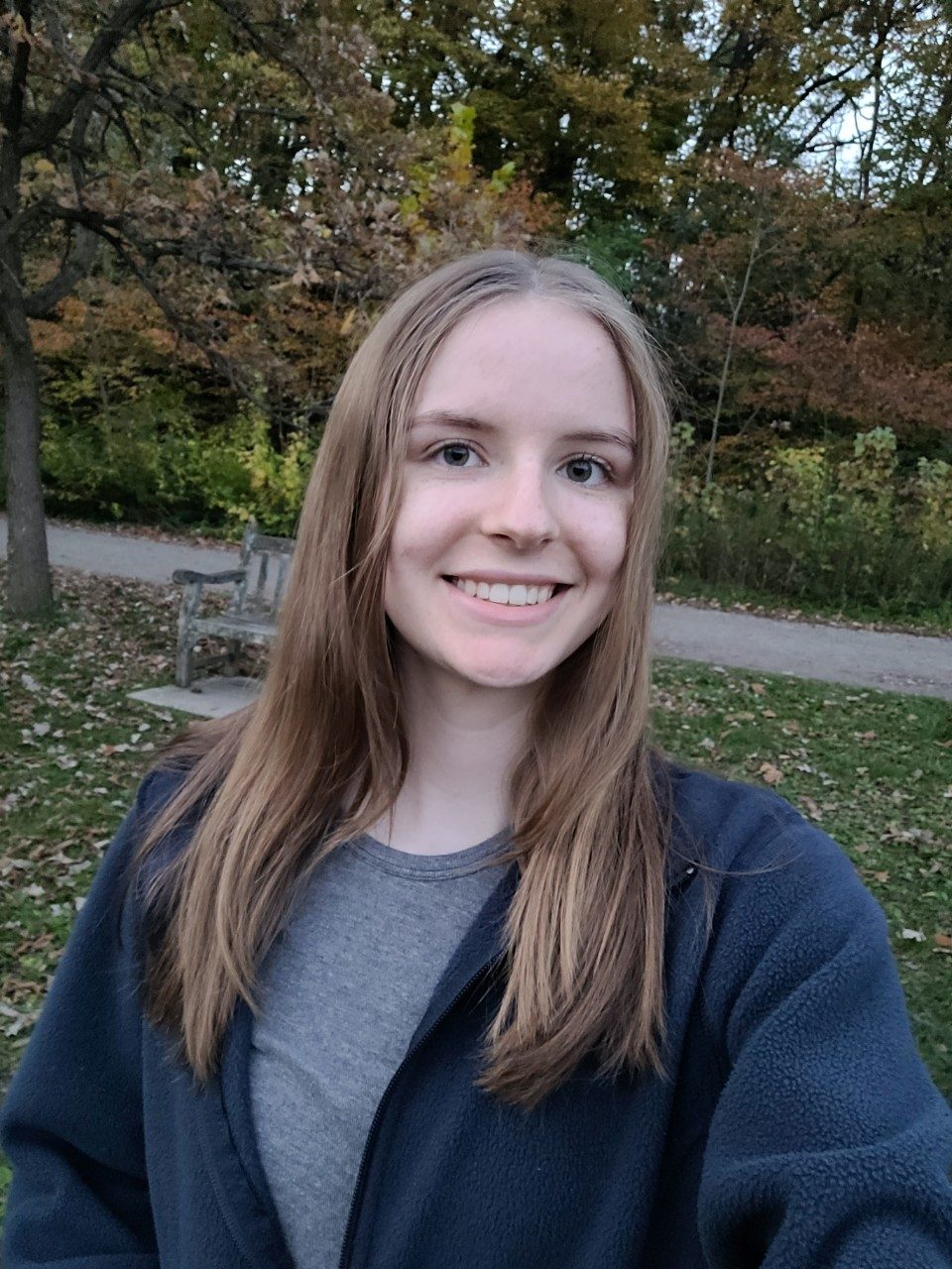 A woman in a grey shirt and blue sweater smiling in front of trees