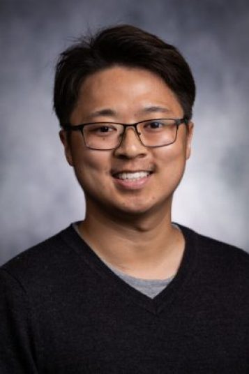 An Asian man with black hair, glasses, and a black shirt smiles at the camera in front of a grey background