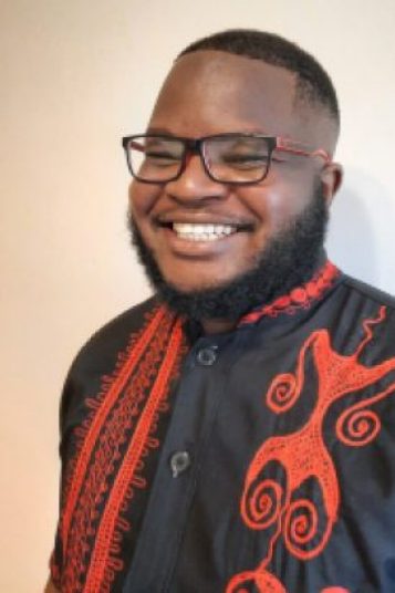 A Black man in glasses and wearing a black and red shirt smiles at the camera. 