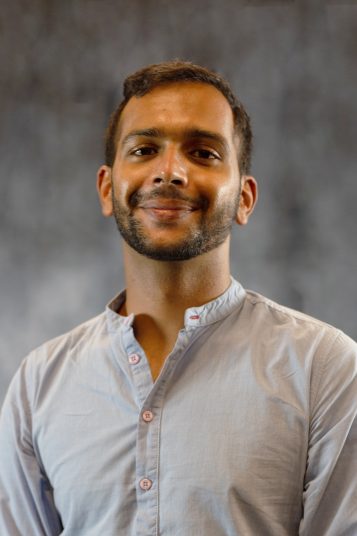 An Indian man with dark hair smiles at the camera, wearing a gray button-up shirt in front of a gray background