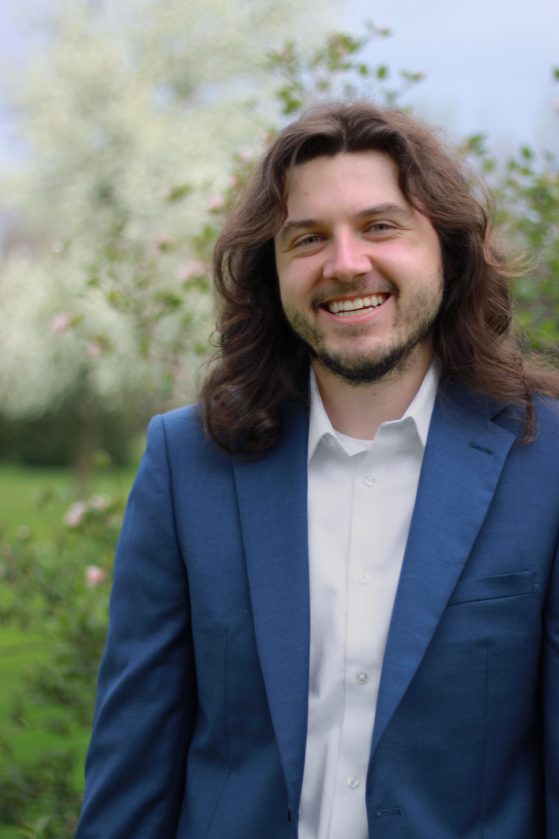A white man with long brown hair smiles to the camera wearing a blue suit coat