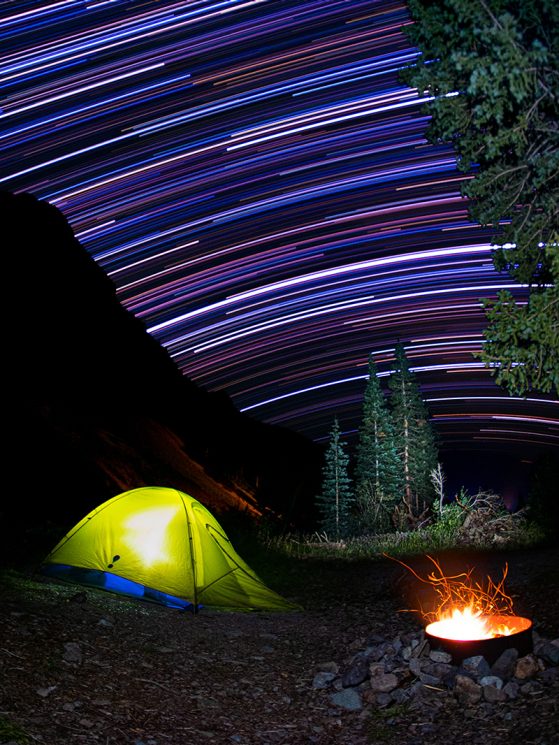 Nightclub, star trails over our campsite in the San Juan mountains of Colorado, Weminuche Wildernes. Image credit: Will Weaver. Extended exposure photography over a campsite with a yellow tent lit up and bonfire, a few trees and a hill, night sky is streaked with star trails of various colors, blue, red, white