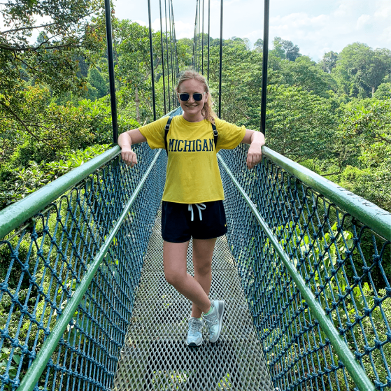 A person is standing on a narrow suspended walking bridge. The bridge appears to be above the treetops of a heavily wooded area. The person is wearing a shirt that reads "Michigan".