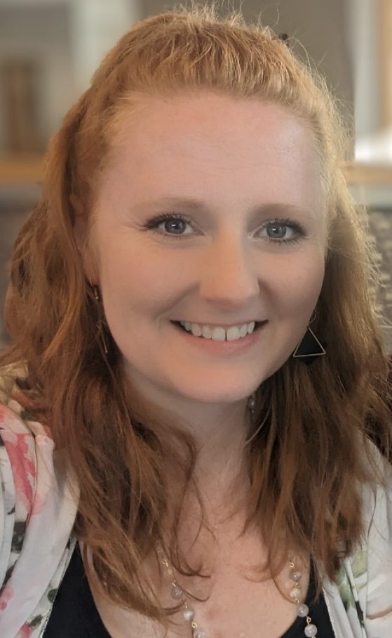 Stephanie is a white woman with long, wavy orange hair and blue eyes. She wears a black top, a sheer kimono with flowers on it, a necklace made of small round stones, dangling gold triangle-shaped earrings and is smiling in front of a blurred background.
