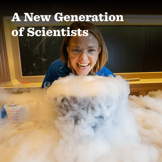 In this photograph, Kate Biberdorf wears a dark blue safety suit and safety goggles and smiles as she stands behind a large bucket on a classroom desk that spews liquid nitrogen all around her. The professor is in a classroom in front of a clean chalkboard. In the foreground this text: A New Generation of Scientists.