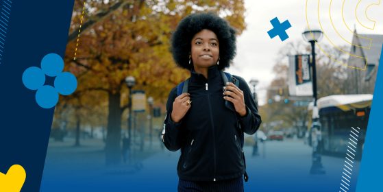 A student is walking on a sidewalk on campus. She is wearing a backpack and is looking head confidently. 