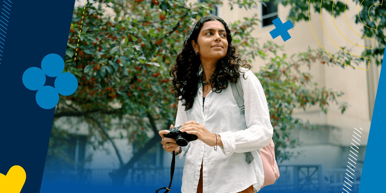 A student on campus holds a camera and looks over her left shoulder. 