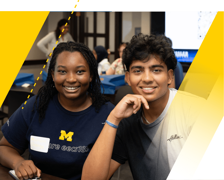 Two students from the Kessler Scholars Program sit at a table. They are facing the camera and smiling. In the background are other people in conversation, sitting and standing, and an illuminated projection screen. 