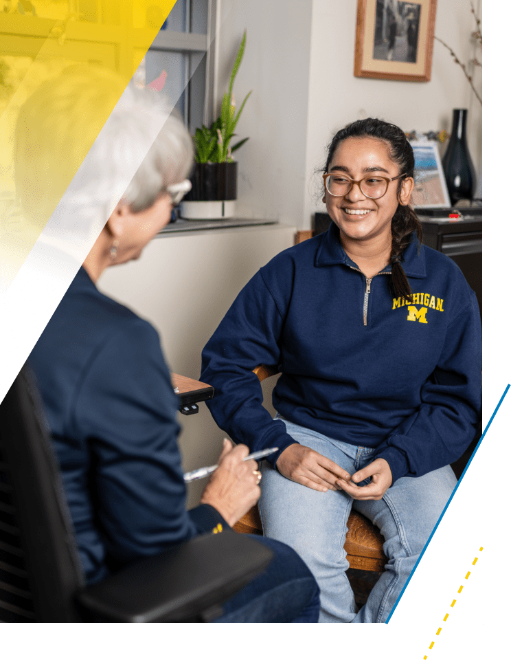 A student meets with a staff member at the Newnan Academic Advising Center. The student sits in a chair, smiling and wearing a navy blue long-sleeve quarter zip shirt with a maize block M on the chest.