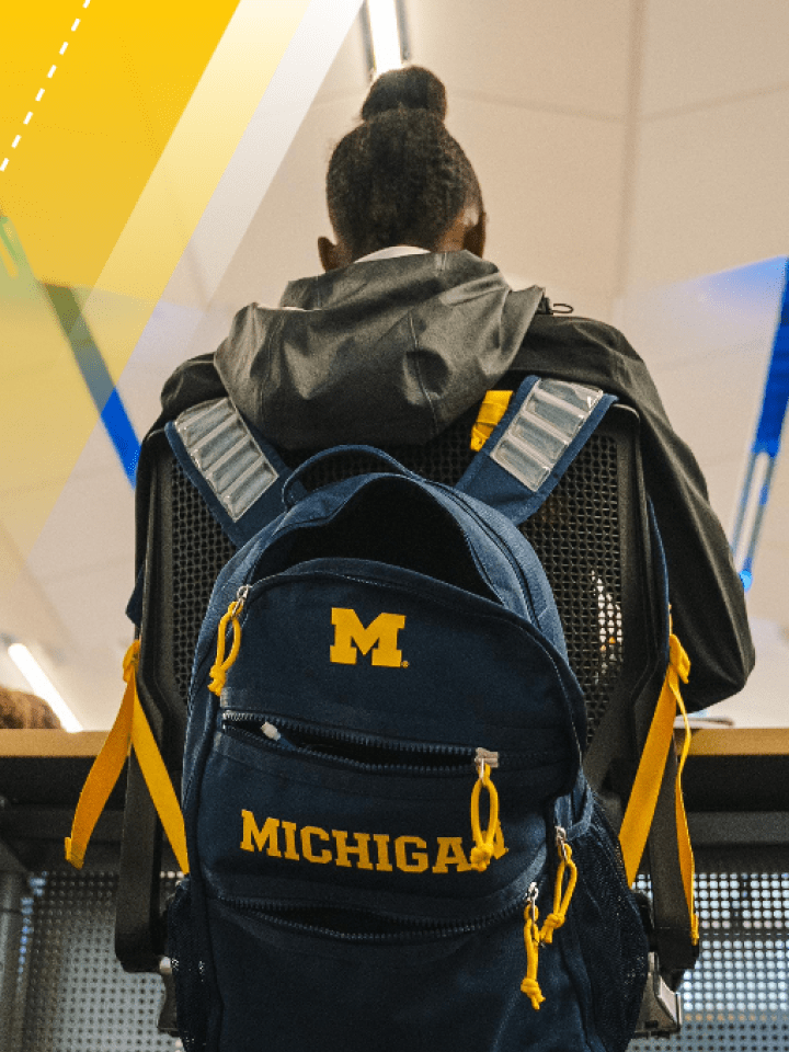 An individual stands at a counter with their back to the camera. They are wearing a blue backpack that features maize details: straps, zipper pulls, a block M, and the word Michigan spelled out in capitalized collegiate font. 