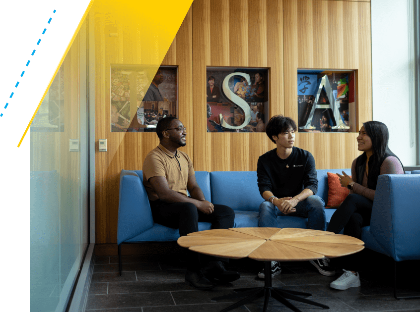 LSA Mental Health and Well-Being Student Advocate Brandon Bond speaks with two individuals on a blue sectional sofa in a lounge area. The letters L, S, and A are decoratively displayed on a light wood-paneled wall behind them. 