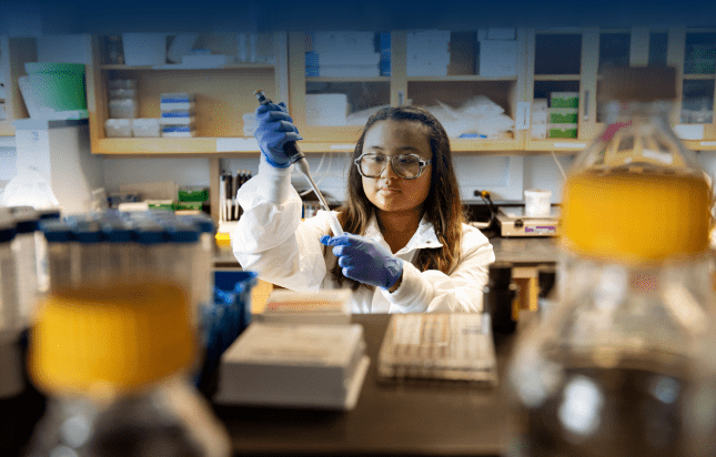 LSA student Clarissa Babila is pictured in Dr. Carl Koschmann’s ground-breaking cancer research lab. Wearing eyeglasses, a white lab coat, blue gloves, and working with a pipette, she is viewed from behind a tall lab counter on which a brightly-capped bottle sits and behind her are shelves and cabinets full of lab items. 