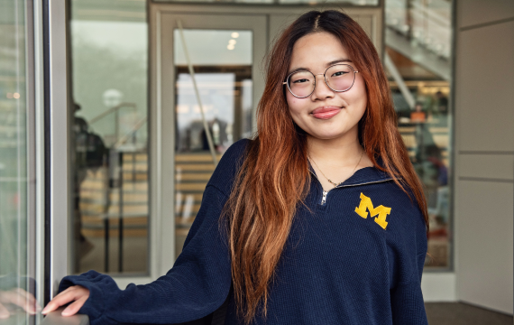 LSA Scholarship student Angelina Wong stands smiling at the entrance to the LSA Building atrium, smiling and wearing a navy blue long-sleeve quarter zip shirt with a maize block M on the chest.