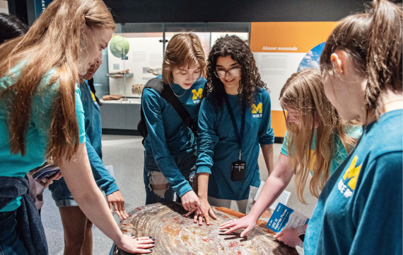 Six students from the Women in Science and Engineering Residence Program (WISE RP) crowd around a hands-on exhibit at a WISE Night at the U-M Museum of Natural History event. They are wearing matching blue WISE RP shirts, in short- and long-sleeved versions.