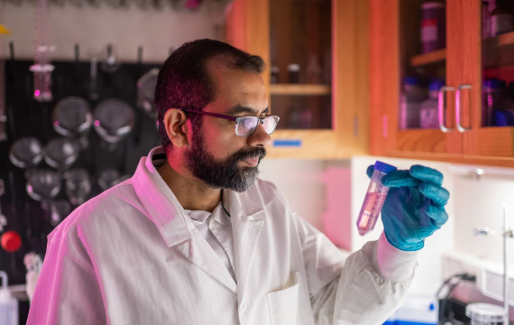 In purple lighting, Professor Omar Ahmed stands in front a lab counter with cabinets above, peering at a test tube filled with liquid. He is wearing eyeglasses, a lab coat and blue gloves. Other lab implements are attached to the rear wall of the room.