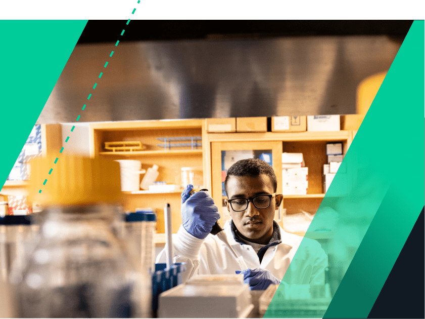 A student wearing eyeglasses, a white lab coat, blue gloves, and working with a pipette is pictured in Dr. Carl Koschmann’s ground-breaking cancer research lab. He is viewed from behind a tall lab counter on which a brightly-capped bottle sits and behind him are shelves and cabinets full of lab items.
