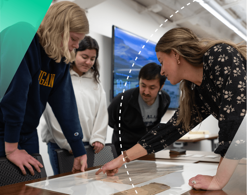 Three students stand looking down at a laminated diagram in Clements Library while a staff member points out something of interest out to them.