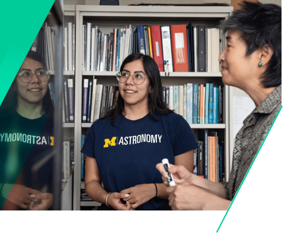 In front of a bookcase in Professor Sally Oey’s office, astronomy Ph.D. student Irene Vargas-Salazar, who has long, straight black hair and glasses, looks at a white board with Oey toward the left side of the image. Both Vargas-Salazar and Oey are smiling. Oey is holding a black EXPO marker and Vargas-Salazar is wearing a U-M Astronomy shirt.