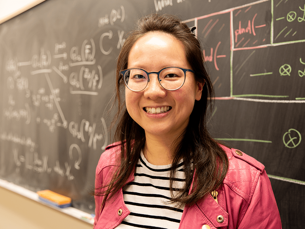 LSA math assistant professor Charlotte Chan in pink sweater and black and white shirt standing in front a chalkboard.