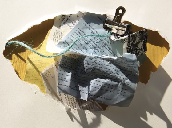 Several fragments of crumpled paper resting in a larger paper fragment. There is a binder clip and a strand of yarn. The writing is not legible.
