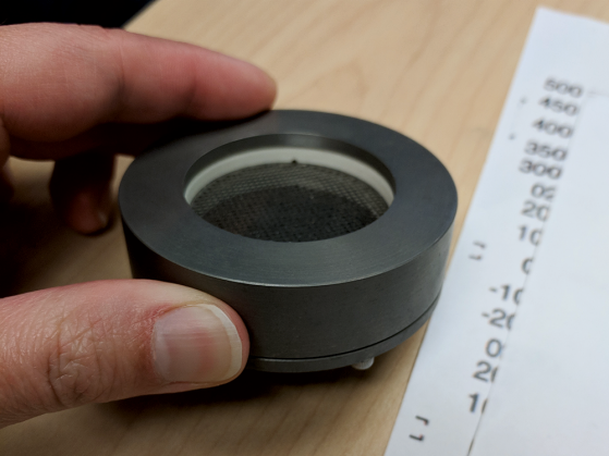 A photograph of an early prototype of the Faraday cup. It's a black round piece that appears hollow in the center. It is positioned between an index finger and a thumb on a table.