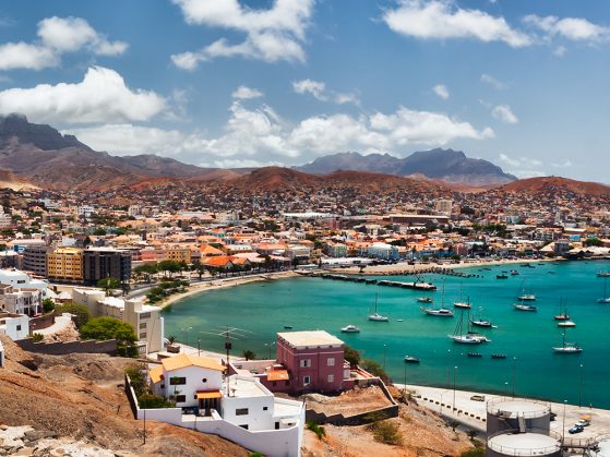 A photograph of a Cape Verdean harbor in which you can see, from a distance, a town bordered by a mountain range, a huge expanse of blue sky with fluffy clouds, and a bright, aqua blue body of water where several sailboats are anchored.