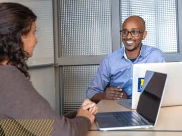 Olivier U. Bahizi, a person with glasses and a blue shirt, meets with a student who has long dark hair. Both are seated facing each other at a desk.