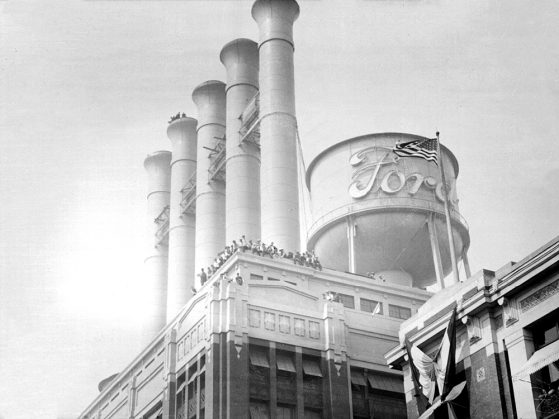 Detail view of the smokestacks and water tower of the power plant at Ford Motor Company Highland Park designed by architect, Albert Kahn, with men standing along top corner of building and one of the smokestacks.