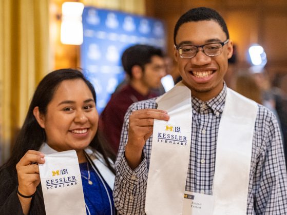 Erica Gonzalez-Paramo and Elijah Taylor smile at the camera. They are each wearing a graduation stole, and they each lift the right side for the camera, which says M|LSA Kessler Scholars