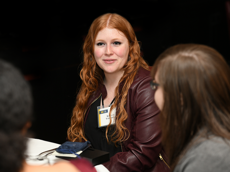 A photograph of Kaitlyn Van Riper sitting at a table with other students. She is wearing a dark red leather jacket with a name tag clipped to the lapel.