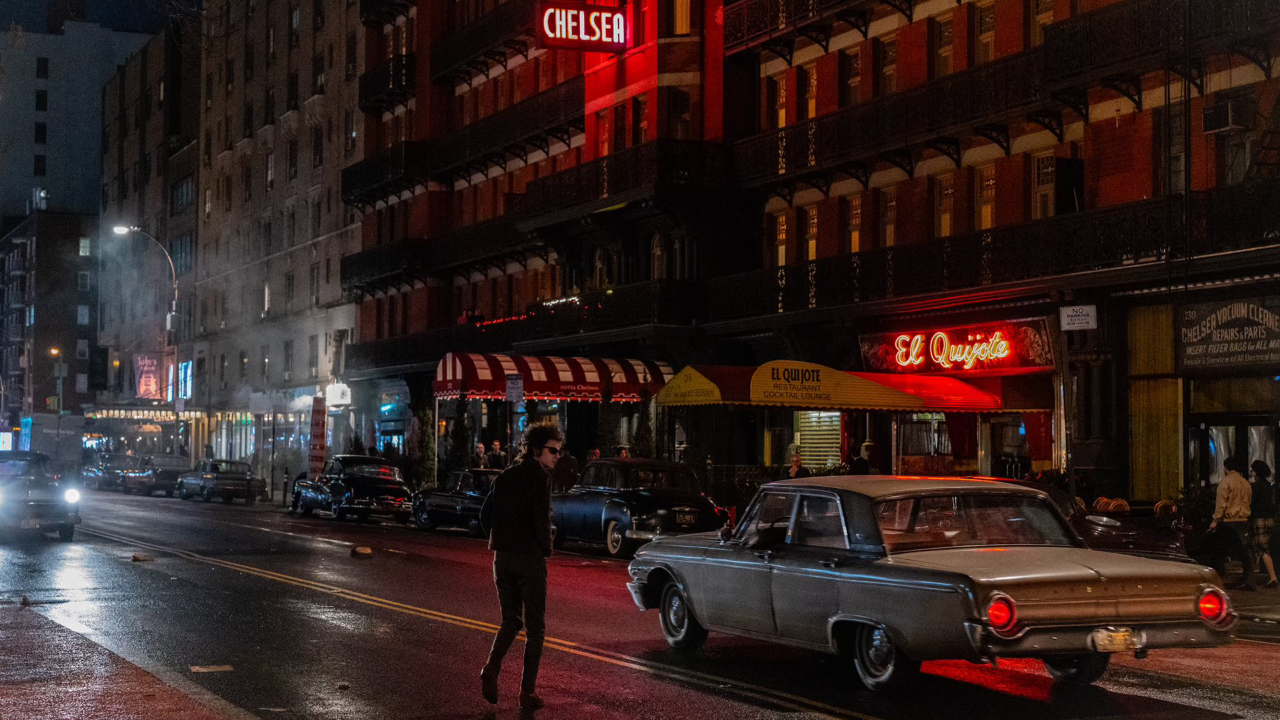 A photograph of a New York City street at night. The actor Timothée Chalamet, in character as Bob Dylan, jaywalks towards the Chelsea Hotel.