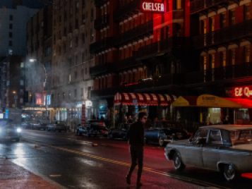 A photograph of a New York City street at night. The actor Timothée Chalamet, in character as Bob Dylan, jaywalks towards the Chelsea Hotel.