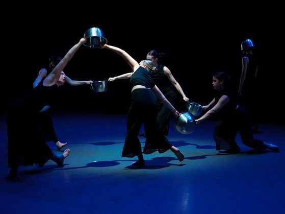 Five dancers wearing all black, holding silver bowls between them, dance in a circle. Another lone dancer stands to their right.