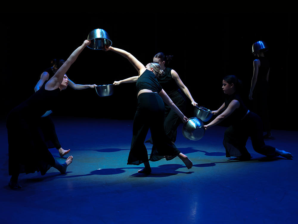 Five dancers wearing all black, holding silver bowls between them, dance in a circle. Another lone dancer stands to their right.