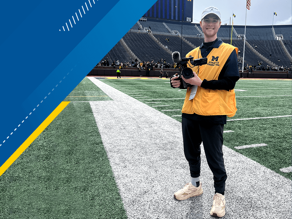 Frank Parkinson wears a maize-colored vest and stands on the field of the Big House holding a camera. 