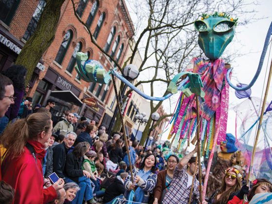 A photograph of a crowd gathered to watch the Festifools parade. A very tall puppet with a green alien head and bug eyes and a body of pink strips supported by sticks rises from the middle of the crowd.