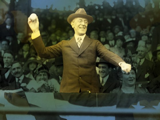A picture of Woodrow Wilson preparing to throw a baseball in front of a crowd. The crowd is tinted blue and Wilson is vibrantly colored -- brown suit and hat, his right arm cocked for a pitch. He has a smile on his face.