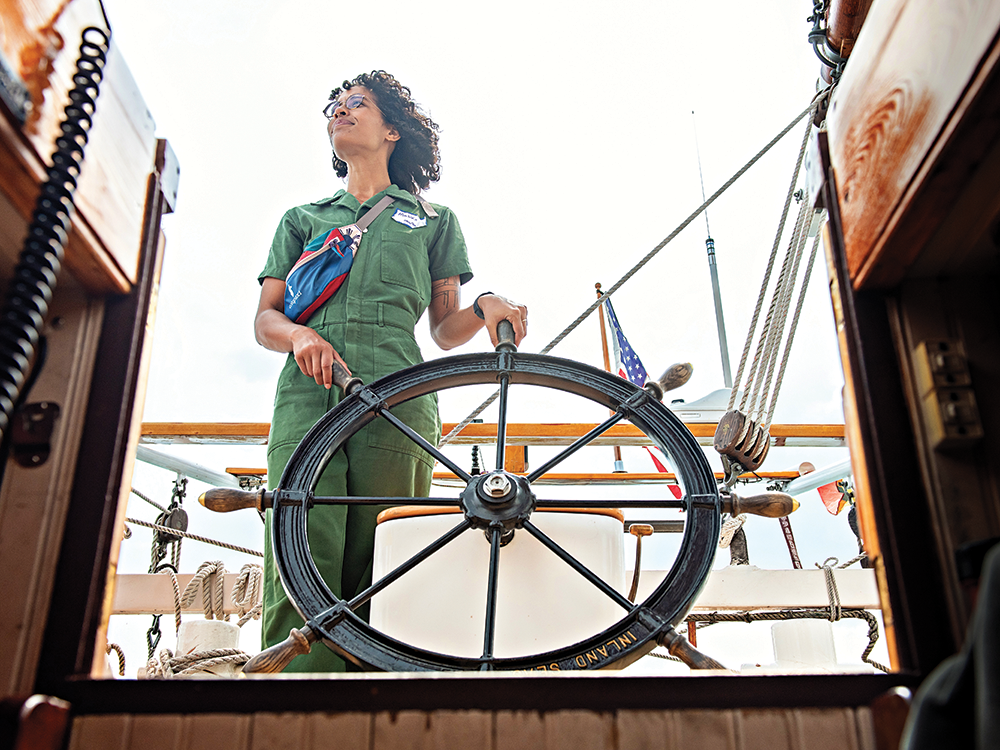 U-M graduate student Malika Stuerznickel stands and steers the Inland Seas schooner on the Detroit River. Her hands are on the steering wheel and she is photographed from the steps leading up from below the deck. She wears a short-sleeved one-piece green jumper and has glasses on.