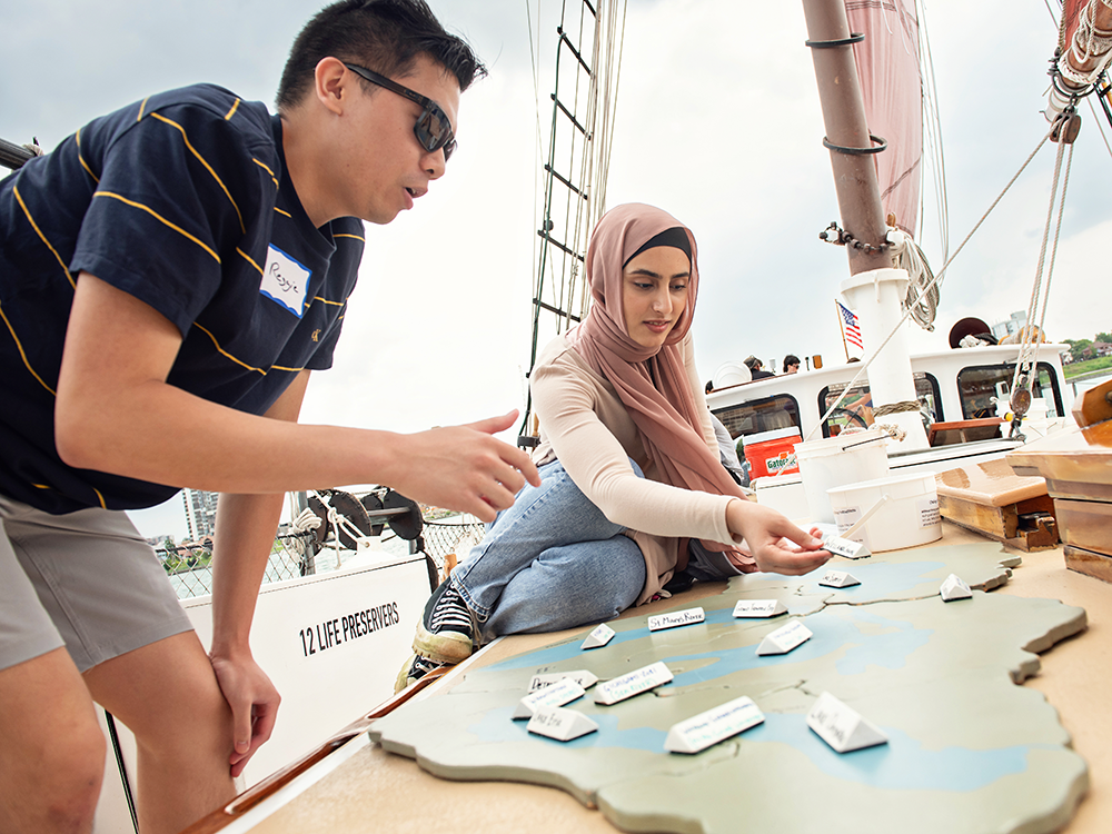 Two U-M students put together a puzzle of the Great Lakes on the deck of the Inland Seas schooner in the Detroit River. One is a male student, who is standing, and one is a female student, who is sitting; she wears a pink head scarf and is placing labels on the top of the puzzle pieces.