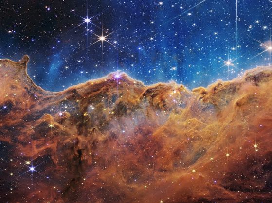 an undulating, translucent star-forming region in the Carina Nebula is shown in this Webb image, hued in ambers and blues; foreground stars with diffraction spikes can be seen, as can a speckling of background points of light through the cloudy nebula