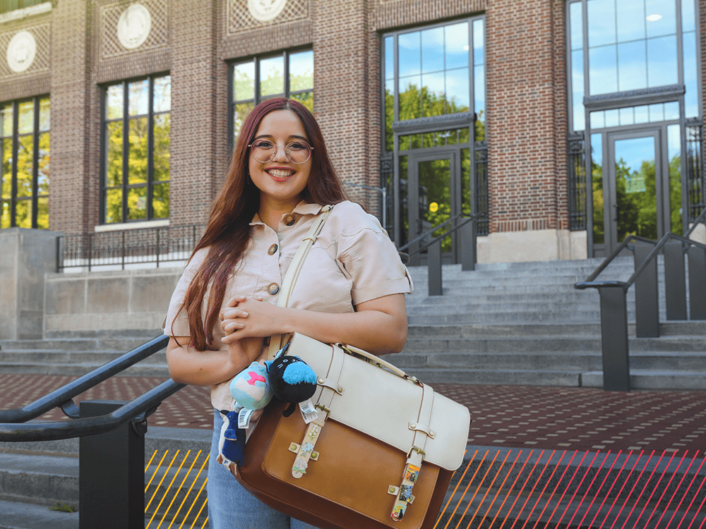 A woman wearing glasses and holding a leather messenger bag smiles directly at the camera.