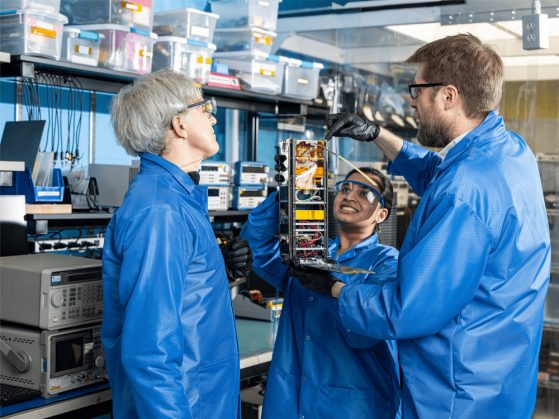 Three researchers in blue jumpsuits examine an object in a science lab.