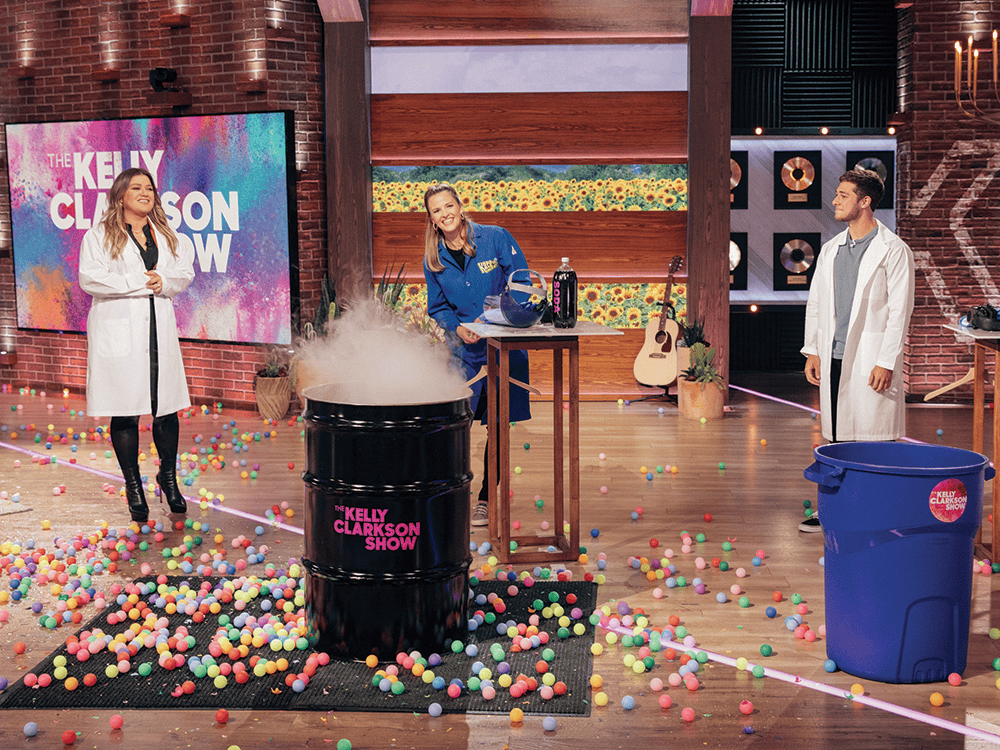In this photograph, Kelly Clarkson stands to the left of her talk show stage in a white lab coat, smiling and looking at Kate Biberdorf who appears behind a large, black trash can with “The Kelly Clarkson Show” written in pink letters on it. Biberdorf is wearing a dark blue safety suit and smiling into the camera. The trash can has liquid nitrogen coming out of the top, and there are different colored balls the size of tennis balls spewed across the stage.